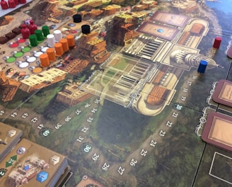 A detailed view of a board game setup with a complex game board featuring a map-like design. Various colored game pieces, including small discs and cylindrical markers in red, green, black, white, and orange, are arranged across the board. There are also decks of cards on the board, each having illustrations and text. The board contains numbered spaces and intricate graphics representing a European city or town. The game appears to be a strategic or historical themed game.