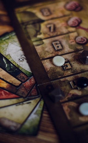 A close-up view of a board game setup, featuring a game board with numbered tiles and various game pieces, including dice and small tokens. Cards in different colors with intricate designs are fanned out beside the board.